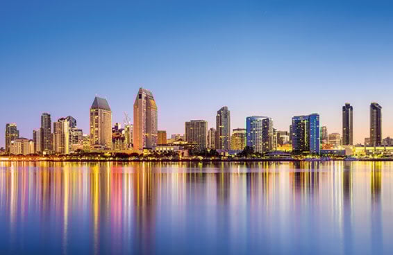 A panoramic view of the San Diego skyline at dusk, with the city reflected in the calm water